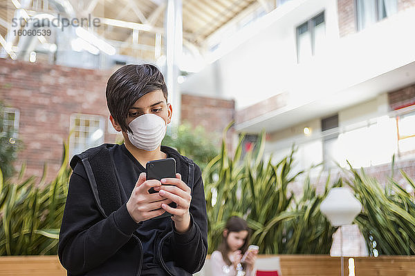 Junge mit Smartphone  der während der Coronavirus-Weltpandemie eine Schutzmaske zum Schutz vor COVID-19 trug  und seine jüngere Schwester im Hintergrund; Toronto  Ontario  Kanada