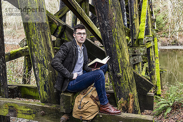 Junger Mann sitzt auf einer moosbewachsenen Bockbrücke neben einem Teich und liest in ruhiger Lage aus einem Buch; Bothell  Washington  Vereinigte Staaten von Amerika