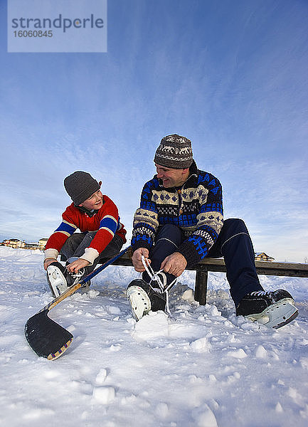 Ein Vater und sein Sohn schnüren ihre Schlittschuhe auf einer Bank auf einem zugefrorenen See für ein Hockeyspiel im Freien; Alberta  Kanada