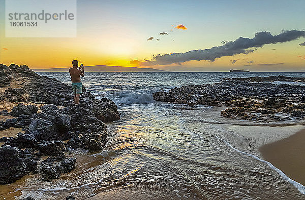 Ein junger Mann im Badeanzug steht auf dem Vulkangestein am Ufer und fängt den goldenen Sonnenuntergang mit einer Smartphone-Kamera ein; Kihei  Maui  Hawaii  Vereinigte Staaten von Amerika