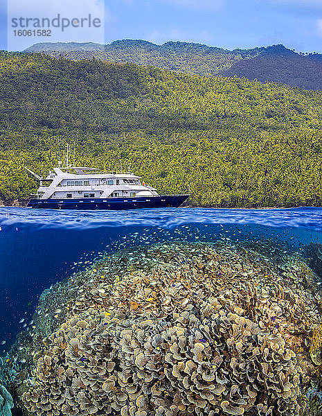 Eine gespaltene Szene mit einem flachen Hartkorallenriff unter Wasser und einer Insel darüber mit dem Live-Bord-Tauchschiff Infiniti; Philippinen