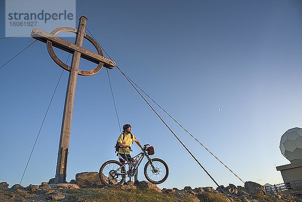 Mountainbiker steht mit e-MTB neben den Gipfelkreuz des Patscherkofel im warmen Abendlicht  Patscherkofel  Patsch  Tirol  Österreich  Europa