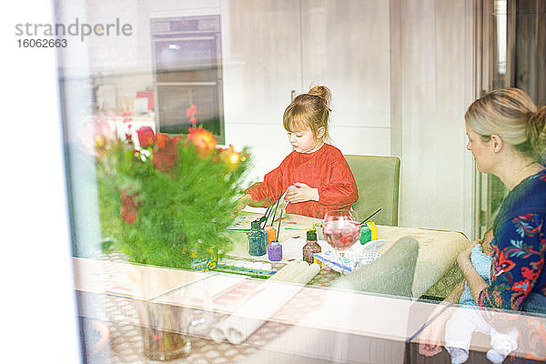 Junges Mädchen benutzt Farben am Küchentisch  während die Mutter in der Nähe sitzt und das Baby hält
