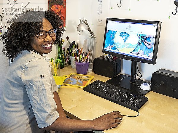 Dunkelhäutige  lächelnde Frau mit Brille arbeitet am Schreibtisch vor Computer  Home Office  Blick in die Kamera  Deutschland  Europa