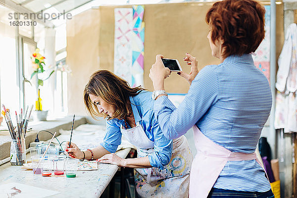 Künstlerin malt im Kreativstudio auf Stoff  reife Frau fotografiert sie mit dem Smartphone