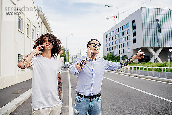 Zwei Männer mit tätowierten Armen stehen auf dem Bürgersteig  benutzen Mobiltelefone und trampen.