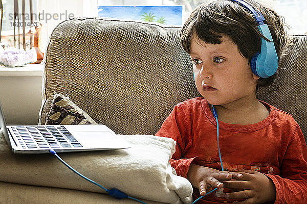 Ein Junge mit braunen Haaren  der blaue Kopfhörer trägt und auf den Bildschirm eines Laptops schaut.