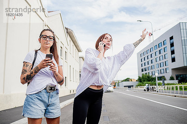 Zwei Frauen mit tätowierten Armen stehen auf dem Bürgersteig  benutzen Mobiltelefone und rufen ein Taxi.