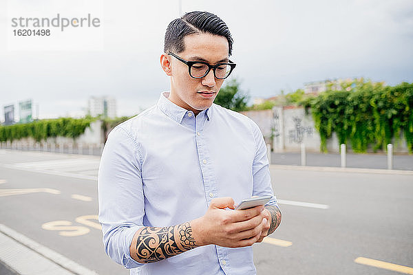 Porträt eines Mannes mit tätowiertem Arm  hellblauem Hemd und Brille  der ein Mobiltelefon benutzt.