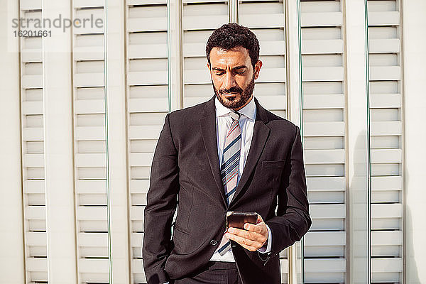 Porträt eines bärtigen Geschäftsmannes in dunklem Anzug  der sein Mobiltelefon überprüft.