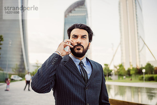 Porträt eines bärtigen Geschäftsmannes in dunklem Anzug  der ein Mobiltelefon benutzt.