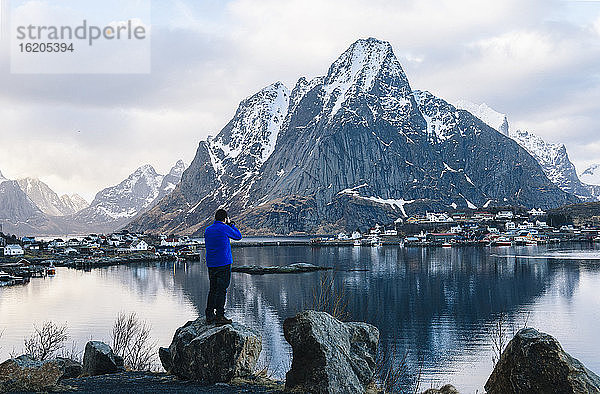 Rückansicht eines männlichen Touristen  der einen schneebedeckten Berg und das Dorf am Wasser fotografiert  Reine  Lofoten  Norwegen