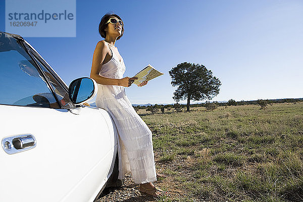 Amerikanische Ureinwohnerin im Sonnenkleid in einem weißen Cabrio-Sportwagen auf unbefestigter Wüstenstraße mit einer Karte