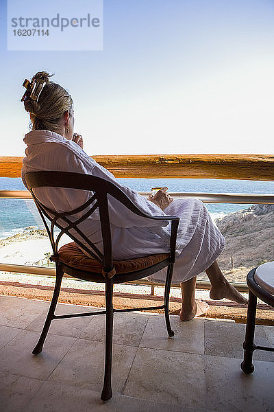 Erwachsene Frau sitzt mit einem Getränk auf einem Hotelbalkon mit Blick auf den blauen Ozean und den weißen Sandstrand