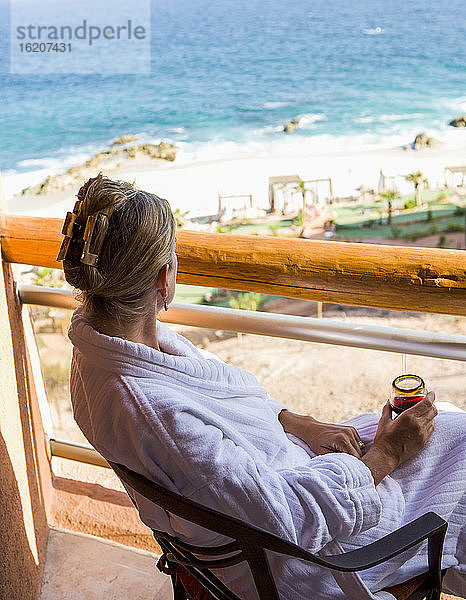 Erwachsene Frau sitzt mit einem Getränk auf einem Hotelbalkon mit Blick auf den blauen Ozean und den weißen Sandstrand