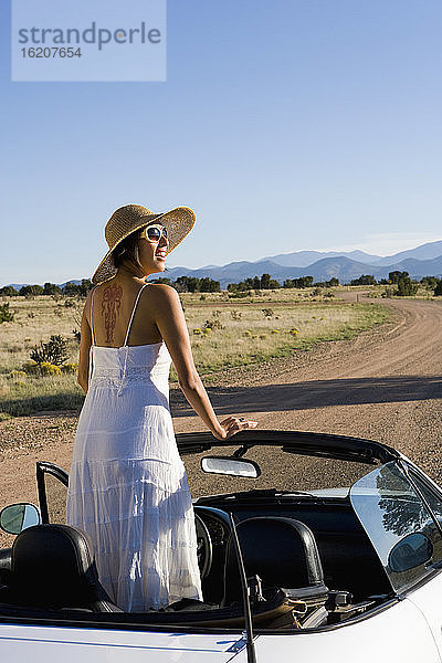 Amerikanische Ureinwohnerin im Sonnenkleid fährt einen weißen Cabrio-Sportwagen auf unbefestigter Wüstenstraße