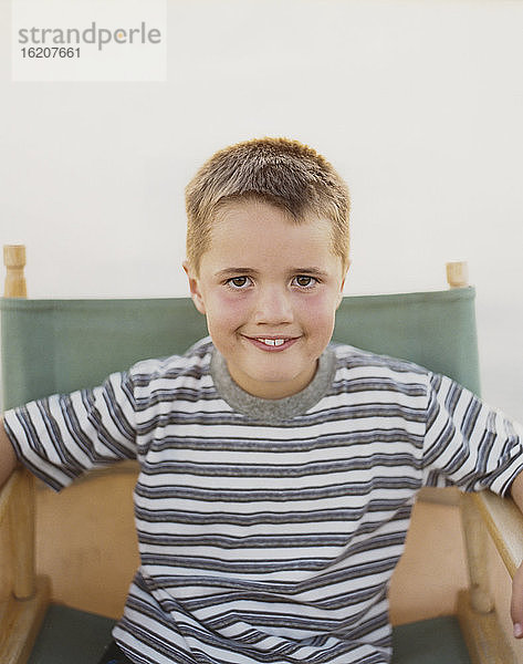 Ein selbstbewusster Junge sitzt auf einem klappbaren Regiestuhl und lächelt mit einem zahnigen Lächeln.