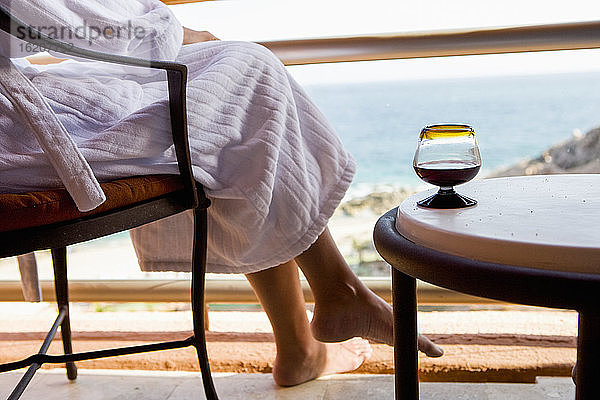 Erwachsene Frau sitzt mit einem Getränk auf einem Hotelbalkon mit Blick auf den blauen Ozean und den weißen Sandstrand