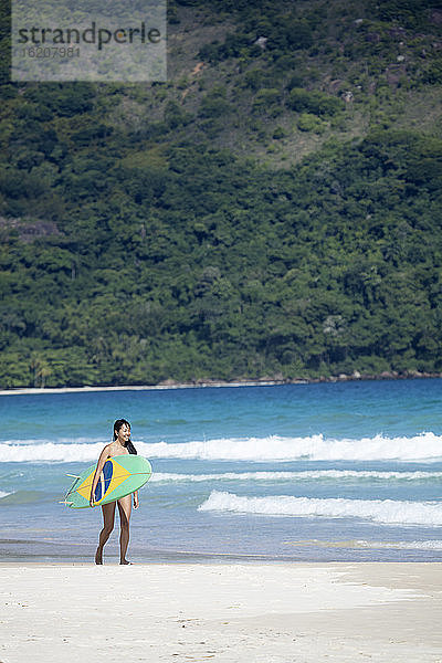 Strandaufnahme eines japanischen Brasilianers (Nipo-brasileiro) im Bikini  der ein mit der brasilianischen Flagge geschmücktes Surfbrett trägt  Brasilien  Südamerika