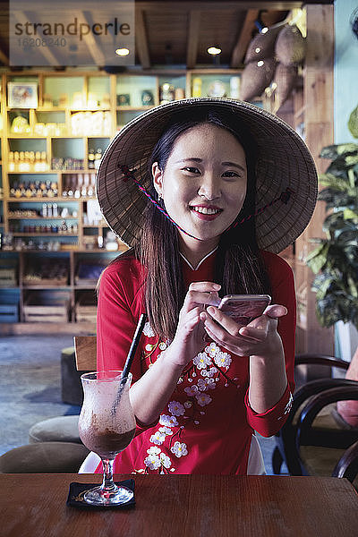 Eine junge Asiatin in rotem Ao Dai-Kleid und konischem Hut lächelt und benutzt ein Mobiltelefon in einem Café  Hoi An  Vietnam  Indochina  Südostasien  Asien