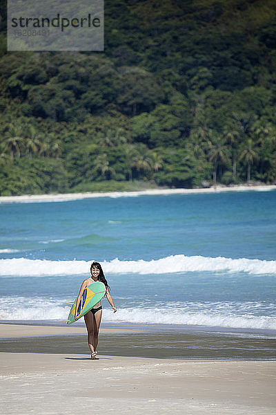 Strandaufnahme eines japanischen Brasilianers (Nipo-brasileiro) im Bikini  der ein mit der brasilianischen Flagge geschmücktes Surfbrett trägt  Brasilien  Südamerika