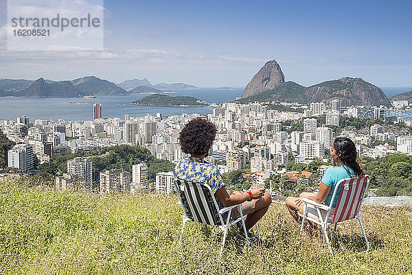 Ein multiethnisches Ehepaar sitzt zusammen und blickt auf den Zuckerhut und die Skyline von Rio  Rio de Janeiro  Brasilien  Südamerika
