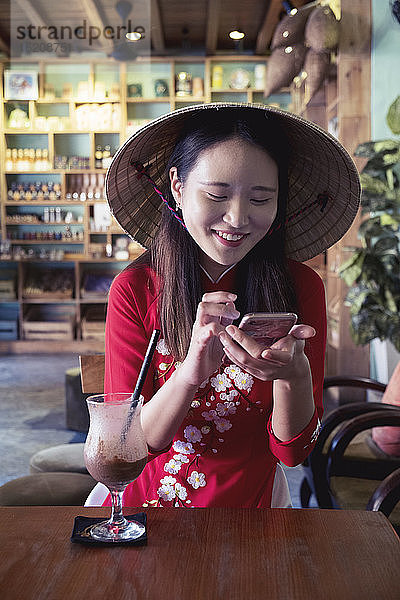 Eine junge Asiatin in rotem Ao Dai-Kleid und konischem Hut lächelt und benutzt ein Mobiltelefon in einem Café  Hoi An  Vietnam  Indochina  Südostasien  Asien