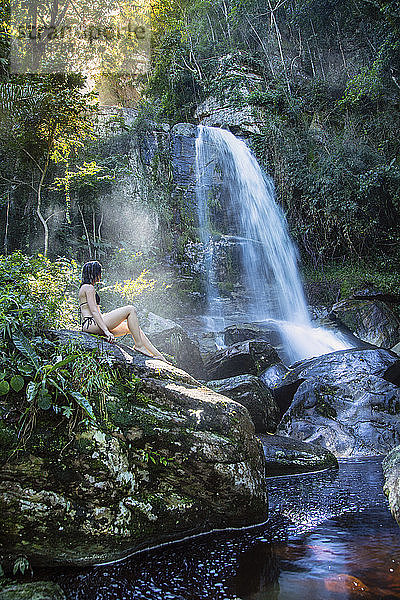 Eine schöne junge Frau sitzt in einem Sonnenstrahl vor einem unberührten Wasserfall im Regenwald  Brasilien  Südamerika