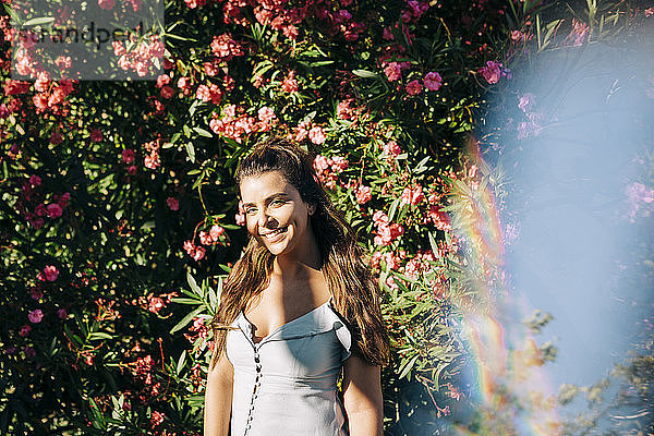 Lächelnde junge Frau mit langen Haaren steht gegen Pflanzen im Park an einem sonnigen Tag