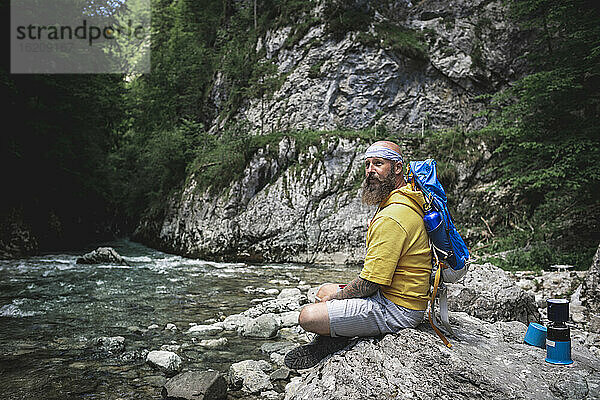Wanderer mit Vollbart und gelbem Kapuzenpulli sitzt auf einem Stein neben einem Fluss