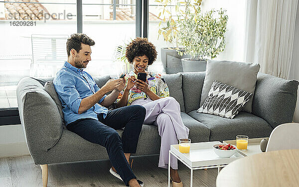 Multiethnisches Paar benutzt Smartphones  während es auf dem Sofa im Penthouse sitzt