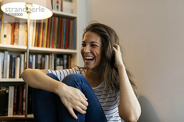 Fröhliche junge Frau mit Hand im Haar  die lachend zu Hause an der Wand sitzt