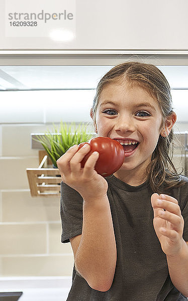 Nahaufnahme eines fröhlichen Mädchens  das eine Tomate in der Hand hält  während es in der Küche zu Hause sitzt