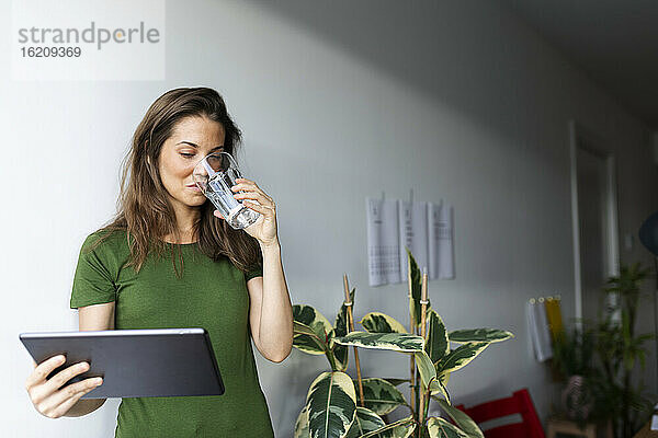 Geschäftsfrau  die Wasser trinkt  während sie ein digitales Tablet an der Wand zu Hause benutzt