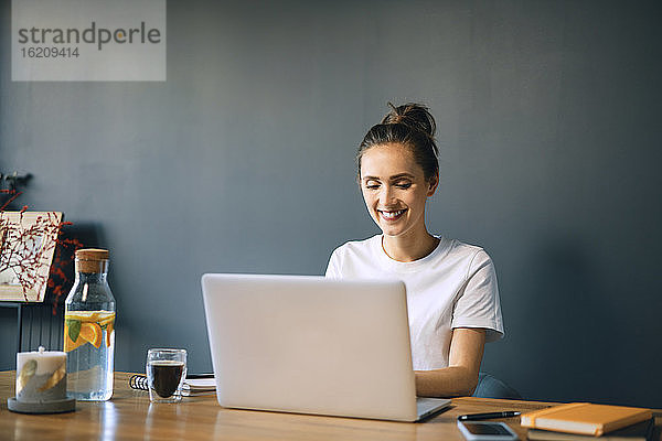 Lächelnde Geschäftsfrau mit Laptop auf dem Schreibtisch an der Wand im Heimbüro