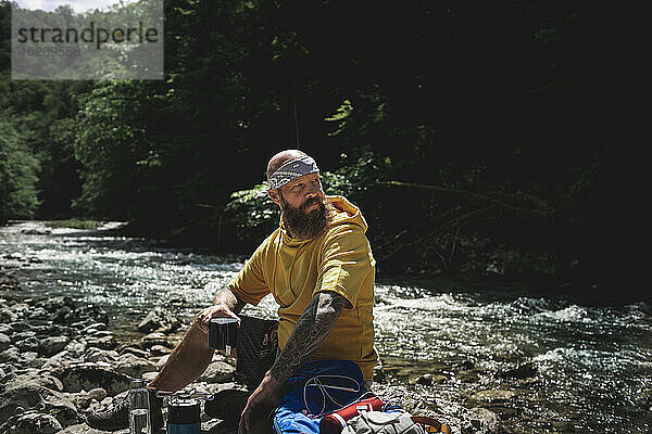 Wanderer mit Vollbart und gelbem Kapuzenpulli bei der Pause  Tee kochen am Flussufer