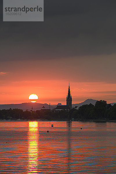 Deutschland  Baden-Württemberg  Radolfzell  Bewölkter Himmel über dem Bodensee bei stimmungsvollem Sonnenuntergang mit Turm des Radolfzeller Münsters im Hintergrund