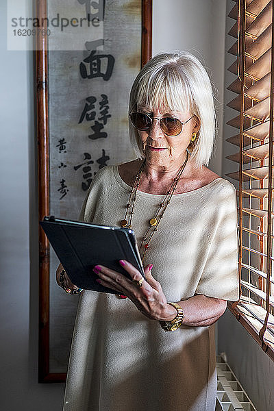 Ältere Frau mit Sonnenbrille  die ein digitales Tablet benutzt  während sie zu Hause am Fenster steht