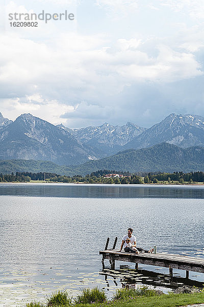 Vater mit Tochter auf dem Steg sitzend über dem See gegen Berge und bewölkten Himmel