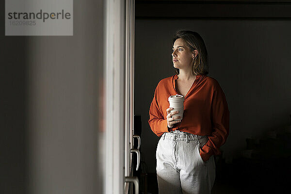 Geschäftsfrau mit Kaffeetasse schaut durch das Fenster zu Hause