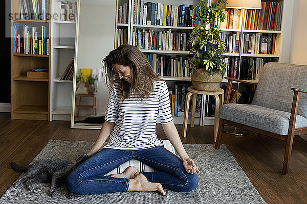 Glückliche junge Frau mit Katze  die zu Hause auf einem Teppich vor einem Bücherregal sitzt
