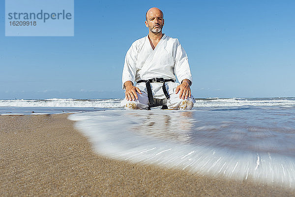 Älterer Mann meditiert kniend am Strand gegen den klaren blauen Himmel