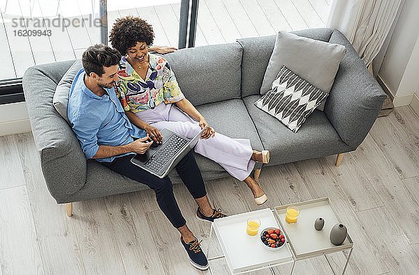Ein multiethnisches Paar benutzt einen Laptop  während es auf dem Sofa im Wohnzimmer eines Penthouses sitzt