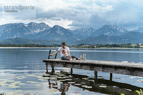 Vater mit Tochter auf einem Steg über dem See vor den Bergen sitzend