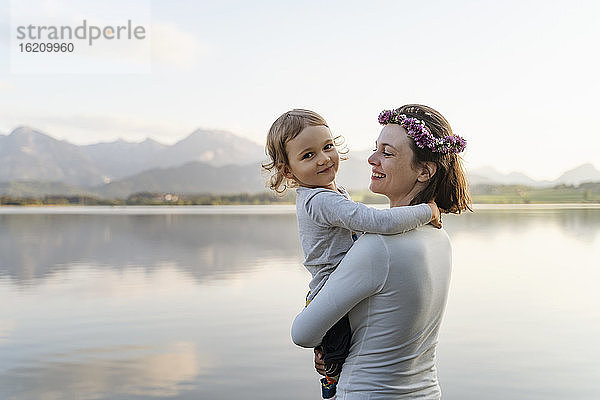 Lächelnde Mutter mit Diadem  die ihre Tochter trägt  während sie bei Sonnenuntergang am See steht