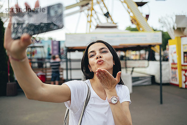 Junge Frau bläst Kuss  während sie ein Selfie im Vergnügungspark macht