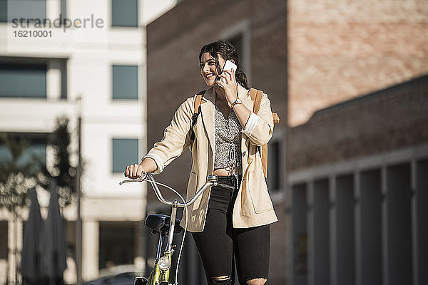 Lächelnde junge Frau  die mit dem Handy telefoniert  während sie mit dem Fahrrad in der Stadt spazieren geht