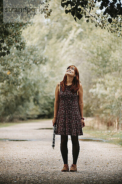 Lächelnde junge Frau  die eine Klarinette in der Hand hält  während sie zu einem Baum hinaufschaut und auf einem Fußweg im Wald steht
