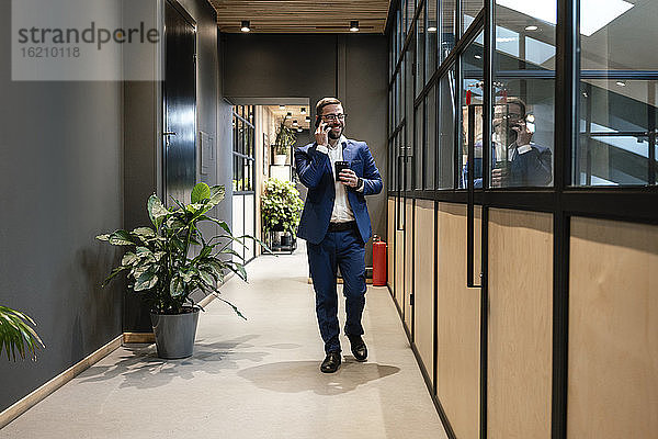 Lächelnder Geschäftsmann  der mit seinem Handy telefoniert  während er eine Tasse an einer Glaswand im Bürokorridor vorbeischiebt