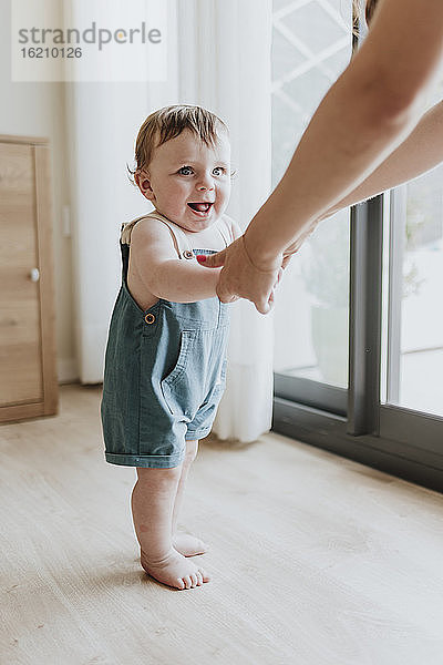 Ein kleines Mädchen hält die Hände seiner Mutter und lernt zu Hause auf dem Boden zu laufen